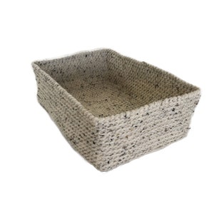 Puede incluir: Una cesta rectangular tejida, de color gris claro con motas negras. La cesta tiene un diseño sencillo y minimalista, probablemente hecha de fibras naturales. La cesta está vacía y sobre un fondo blanco.