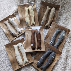 Könnte beinhalten: Set aus Filzfisch-Anhängern in verschiedenen Farben, darunter Grau, Braun und Weiß. Jeder Fisch ist handgenäht und hat ein kleines Pluszeichen an der Seite. Die Anhänger sind auf braunen Karten mit einem Blumenmuster und dem Text "Gefüllt mit Schafwolle" verpackt.