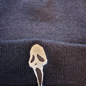 Peut inclure: Un bonnet en tricot bleu marine foncé avec un masque Ghostface blanc et noir brodé, issu de la franchise Scream. Le bonnet est fait d'une matière douce et texturée, adapté au froid.