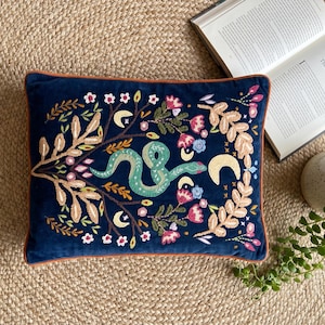 Puede incluir: Un cojín rectangular de terciopelo azul marino con un intrincado bordado de una serpiente verde centrada entre ramas florales estilizadas, hojas y lunas crecientes en tonos rosa, amarillo, blanco y beige, con un borde de ribete naranja.