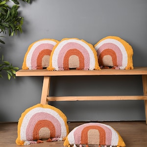 May include: Five rainbow-shaped throw pillows with a white, pink, orange, and yellow color scheme. The pillows have tassels on the bottom and are arranged on a wooden bench and on the floor.