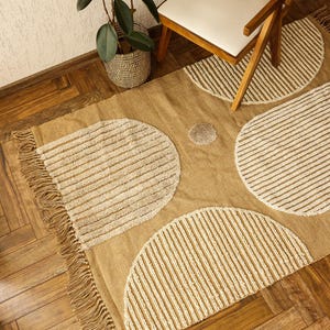 Puede incluir: Una alfombra tejida beige y blanca con borde de flecos presenta un patrón geométrico de tres formas semicirculares grandes. La alfombra está sobre un suelo de madera dura.