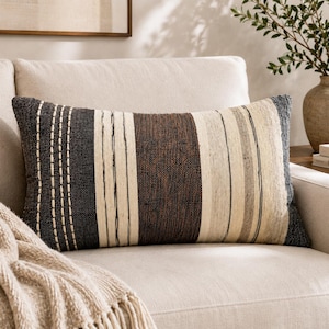 Puede incluir: Un cojín rectangular con textura tejida y rayas verticales en tonos negro carbón, marrón oscuro, crema y gris claro. El diseño incluye detalles decorativos de costuras blancas sobre la sección de color negro.