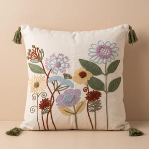 Puede incluir: Un cojín cuadrado de color blanco roto con flores silvestres bordadas en tonos lavanda, amarillo, óxido y azul. El cojín está decorado con pequeñas borlas verdes en cada una de las cuatro esquinas, añadiendo textura al diseño floral.