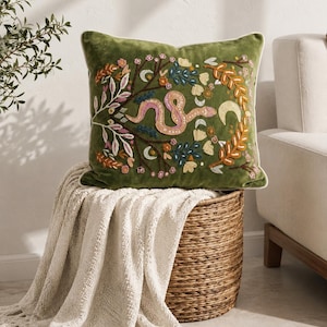 May include: A square olive green velvet throw pillow featuring intricate embroidery of a central pink snake surrounded by colorful floral branches, leaves, and crescent moon motifs, finished with a cream-colored piped edge trim.