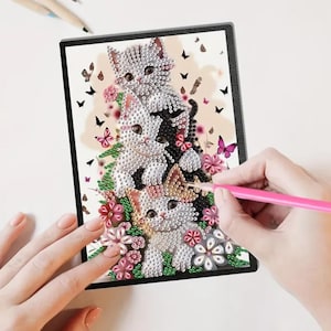 May include: A diamond painting kit featuring a design of three kittens surrounded by flowers and butterflies. The artwork is in a black frame, with a pink stylus held over the canvas. The kittens are white, gray, and black.
