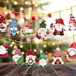 May include: A collection of Christmas gnome ornaments, each with a unique design. The gnomes feature red, green, and white hats, beards, and festive outfits. They are strung on a gold cord against a blurred Christmas tree background.
