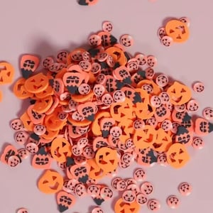 May include: A collection of orange and pink Halloween-themed confetti in various shapes, including jack-o'-lanterns and small circles. The confetti is scattered on a light pink surface, creating a festive and decorative display.