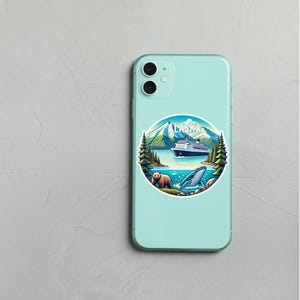 May include: A light mint green smartphone case featuring a circular sticker with a graphic of Alaska. The design includes snow-capped mountains, a cruise ship, evergreen trees, a brown bear, and a whale in blue water with the text ALASKA.