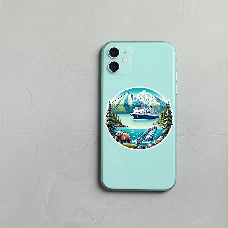 May include: A light mint green smartphone case featuring a circular sticker with a graphic of Alaska. The design includes snow-capped mountains, a cruise ship, evergreen trees, a brown bear, and a whale in blue water with the text ALASKA.