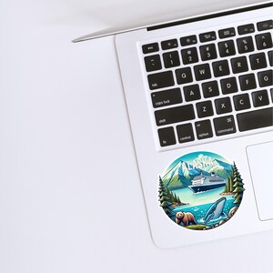 May include: A circular sticker featuring a colorful illustration of an Alaskan landscape with a cruise ship, mountains, pine trees, a brown bear, and a whale. The word ALASKA is printed in white at the top of the sticker, which is placed on a laptop.