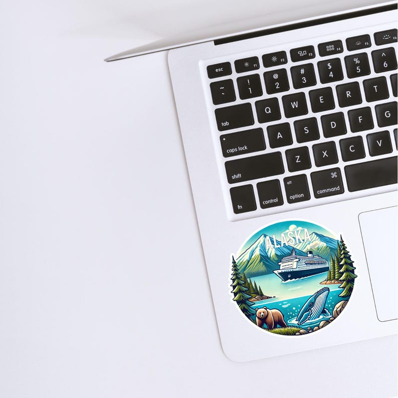May include: A circular sticker featuring a colorful illustration of an Alaskan landscape with a cruise ship, mountains, pine trees, a brown bear, and a whale. The word ALASKA is printed in white at the top of the sticker, which is placed on a laptop.