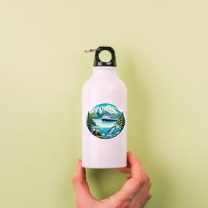 May include: A white metal water bottle with a black cap and a circular sticker featuring a landscape illustration of Alaska. The graphic depicts a cruise ship on blue water, green pine trees, snow-capped mountains, a brown bear, and a whale.