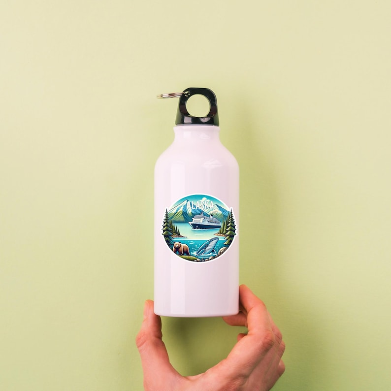 May include: A white metal water bottle with a black cap and a circular sticker featuring a landscape illustration of Alaska. The graphic depicts a cruise ship on blue water, green pine trees, snow-capped mountains, a brown bear, and a whale.