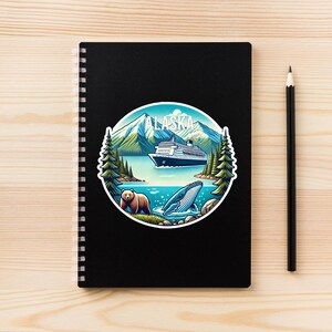 May include: A black spiral-bound notebook featuring a circular sticker with an Alaska landscape illustration. The design includes snow-capped mountains, a cruise ship, evergreen trees, a brown bear, and a whale, with the text ALASKA at the top.