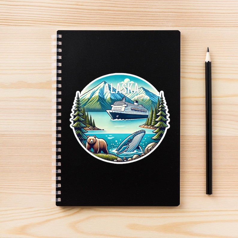 May include: A black spiral-bound notebook featuring a circular sticker with an Alaska landscape illustration. The design includes snow-capped mountains, a cruise ship, evergreen trees, a brown bear, and a whale, with the text ALASKA at the top.