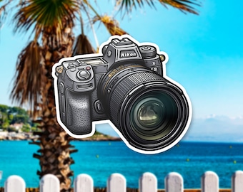 May include: A sticker of a Nikon camera with a large lens, set against a tropical beach scene. The camera is detailed in shades of gray and black, with the Nikon logo visible. The background features a palm tree, blue sky, and ocean.