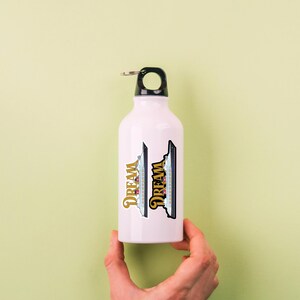Puede incluir: Una botella de agua blanca con una tapa negra y un lazo plateado. La botella tiene dos pegatinas con un dise&ntilde;o de crucero y la palabra "DREAM" en escritura dorada. Sobre un fondo verde claro.