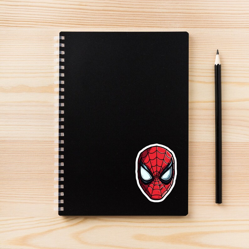 May include: A black spiral notebook with a red and black Spiderman sticker on the front. A black pencil is next to the notebook on a wooden surface.