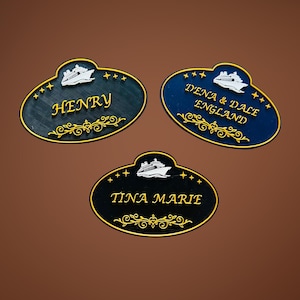 May include: Three oval-shaped name tags with a cruise ship design. Each tag has a different name in gold lettering: "Henry", "Dena & Dale England", and "Tina Marie". The tags are dark blue, teal, and black with gold trim and star accents.