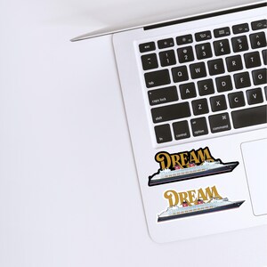 Puede incluir: Dos pegatinas con un dise&ntilde;o de crucero y la palabra "DREAM" en letras doradas. Las pegatinas est&aacute;n colocadas en un port&aacute;til blanco, con el teclado visible. El crucero tiene detalles en rojo y blanco.