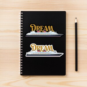 Puede incluir: Dos pegatinas con un dise&ntilde;o de crucero con la palabra "DREAM" en letras doradas. El barco es blanco con un casco negro, chimeneas rojas y detalles azules. Las pegatinas est&aacute;n en un cuaderno negro.