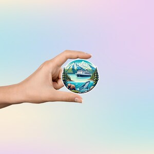 May include: A circular sticker featuring a colorful illustration of an Alaskan landscape with a cruise ship on the water, a brown bear on the shore, a whale breaching in the foreground, tall pine trees, and mountains under the text ALASKA.