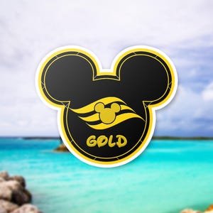 May include: A black and gold Mickey Mouse-shaped sticker with the word "GOLD" at the bottom. The sticker features a yellow Mickey Mouse head and wave design. The background is a blurred image of a beach and ocean.