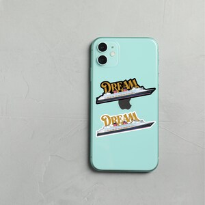 Puede incluir: Un iPhone 11 Pro azul claro con dos pegatinas de un crucero y la palabra "DREAM" en letras doradas. Las pegatinas son en blanco y negro con detalles en rojo y dorado. El tel&eacute;fono est&aacute; sobre una superficie gris claro.