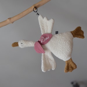 Könnte beinhalten: Ein weißes Plüsch-Gänse-Spielzeug mit braunen Füßen und einem rosa Stoffschal mit dem Namen "Romi" bestickt. Die Gans hängt an einem Ast.