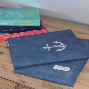 Könnte beinhalten: Zwei blaue Stoffhüllen für Laptops mit einem gestickten Ankerdesign auf der Vorderseite. Die obere Hülle hat ein weißes Ankerdesign. Die untere Hülle hat ein weißes Etikett mit dem Text "Handmade with Love" darauf.