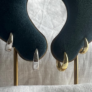 May include: Two pairs of earrings, one silver and one gold, presented on a dark teal velvet surface. The earrings feature a simple, curved design with a polished finish. The gold earrings have a warm, shiny appearance, while the silver earrings have a bright, reflective surface.