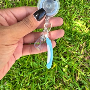 May include: A clear circular retractable badge reel featuring a metal spring mechanism and a transparent plastic strap. Attached to the strap is a small, curved, light blue and white charm shaped like a disposable razor with a textured handle.