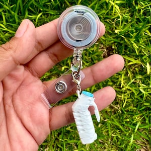 May include: A clear plastic retractable badge reel featuring a circular housing with an internal metal spring mechanism. Attached to the reel is a clear vinyl strap with a silver metal snap button and a white plastic jug charm with a blue cap.