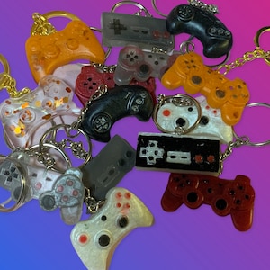 May include: A collection of colorful, resin-cast video game controller keychains. The keychains feature different designs, including classic controllers, modern controllers, and even a retro NES controller.