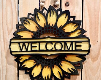 May include: A wooden welcome sign with a black and yellow sunflower design. The sign says "WELCOME" in black letters.