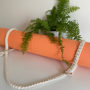 May include: A white crocheted yoga mat strap with a ring on one end. The strap is wrapped around an orange yoga mat. A green fern plant is in the background.