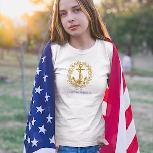May include: White t-shirt with a gold anchor design inside a floral wreath. The shirt also features the text "N.O.B" and coordinates. The person is wearing blue jeans and draped with an American flag.