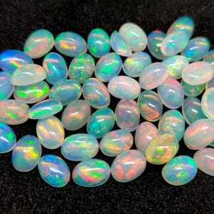 Könnte beinhalten: Eine Ansammlung ovalförmiger Opale mit schillernden Farben wie Blau, Grün und Orange. Die polierten Opale zeigen ein Lichtspiel auf ihren Oberflächen. Die Edelsteine sind vor einem dunklen Hintergrund verstreut.