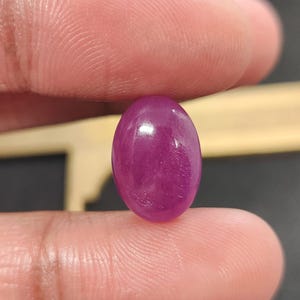May include: A polished, oval-shaped ruby gemstone. The gem is a deep, saturated magenta color with a smooth, reflective surface. The ruby is held between two fingers, showcasing its size and shape.