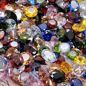 May include: A collection of colorful gemstones in various shapes and sizes. The gems are in shades of red, pink, yellow, blue, green, and clear. The gemstones are faceted and reflect light, creating a sparkling effect.