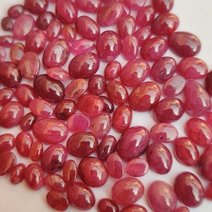 May include: A collection of oval-shaped, polished gemstones in varying shades of red and pink. The gemstones are scattered across a white surface, with light reflecting off their smooth surfaces. The gems vary in size.