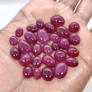 Puede incluir: Una colección de piedras preciosas de rubí pulidas, de forma ovalada y de varios tamaños. Los rubíes muestran un color rojo intenso y rico con un acabado brillante. Las piedras preciosas se sostienen en una mano, mostrando su belleza natural.