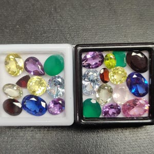 Puede incluir: Dos pequeñas bandejas blancas y negras llenas de varias piedras preciosas de colores. Las piedras preciosas son en tonos de amarillo, morado, azul, verde y rojo. Las piedras preciosas son ovaladas, redondas y en forma de pera.