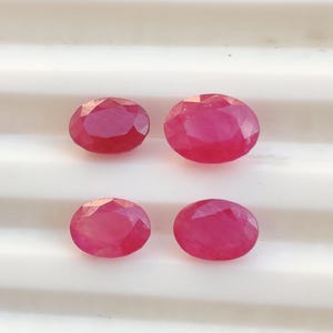 Könnte beinhalten: Vier oval geschliffene Rubine mit einem leuchtenden pink-roten Farbton. Jeder Edelstein hat eine polierte Oberfläche mit mehreren geometrischen Facetten, die das Licht reflektieren, angeordnet in einem zwei-mal-zwei Raster auf einer weißen, gerillten Oberfläche.