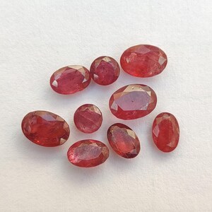 Puede incluir: Una colección de ocho piedras preciosas rojas facetadas en varias formas ovaladas y redondas. Cada piedra muestra un tono rojo vibrante y profundo con facetas geométricas intrincadas que reflejan la luz, exhibiendo una apariencia pulida y translúcida.