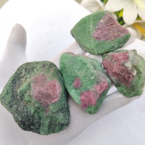 May include: Four rough-cut ruby in zoisite stones. The stones are a mix of green and dark pink. The stones are held in a white gloved hand. The stones are natural and have a rough texture.