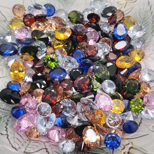 May include: A collection of assorted gemstones in various colors, including blue, yellow, green, pink, and clear. The gemstones are oval and round cut, with a sparkling, reflective surface. The gems are scattered in a clear glass bowl.