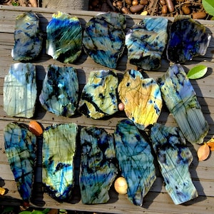 May include: A selection of iridescent labradorite stones, showcasing blues, greens, and yellows. The rough-cut gemstones have a natural, irregular shape, with visible textures and patterns. The stones are arranged on a wooden surface.