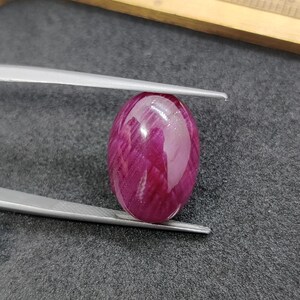 May include: A polished, oval-shaped ruby gemstone, held by tweezers. The ruby displays a deep, rich red color with internal veining, and a glossy surface. The gemstone is set against a dark gray background.